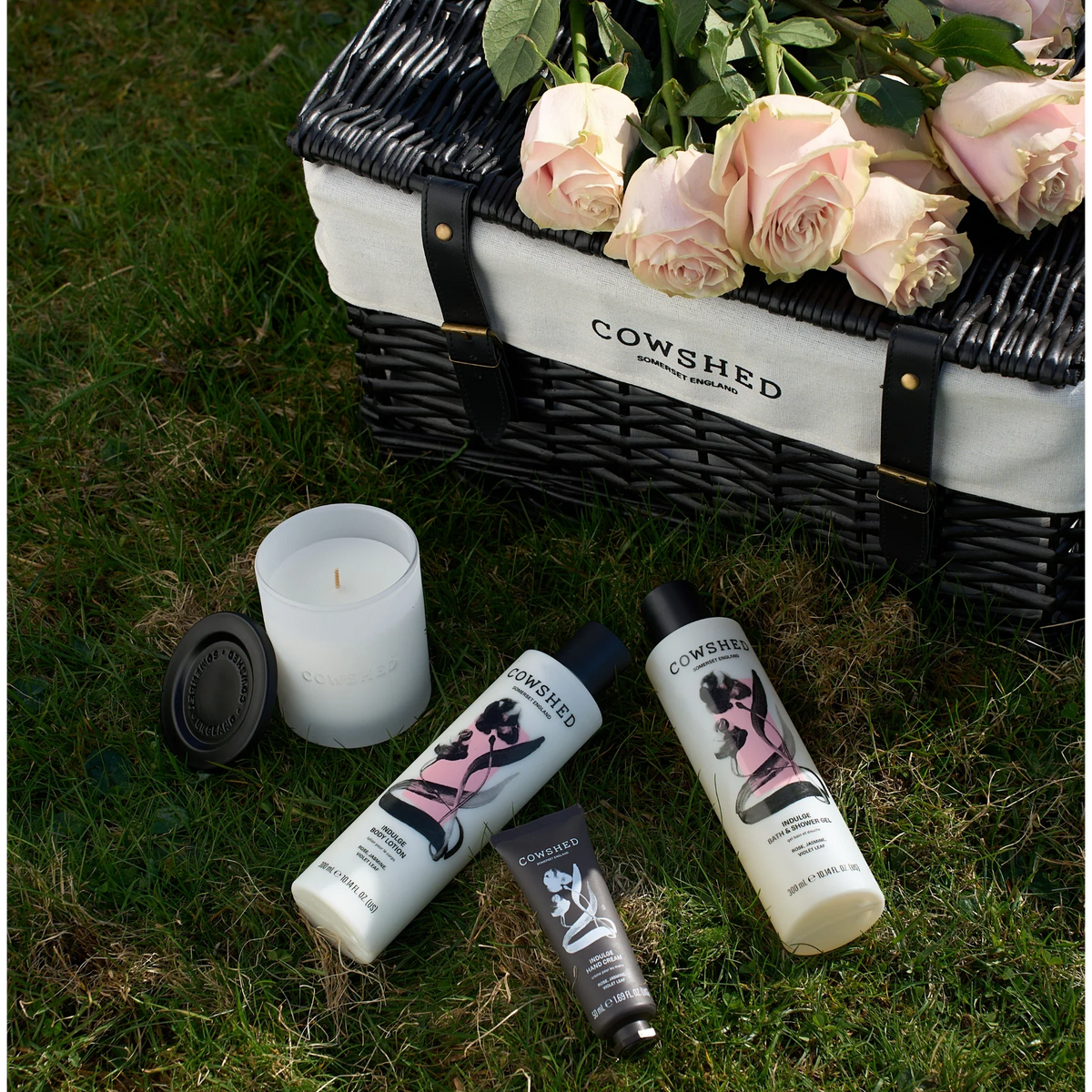 Cowshed Indulge range on the grass next to a hamper and roses