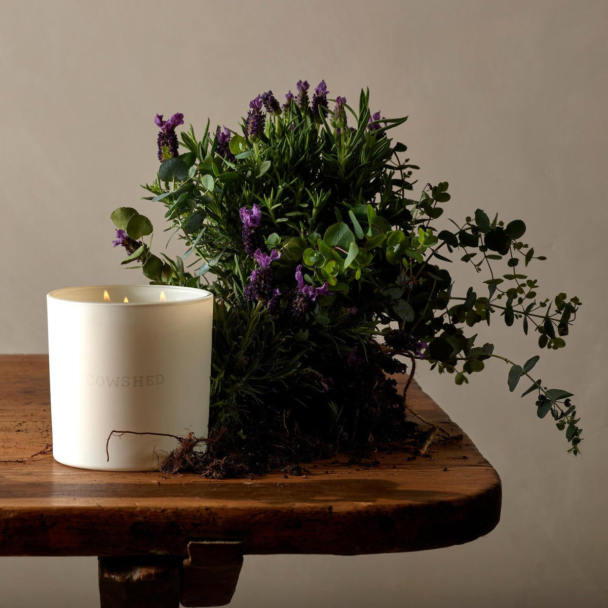 White candle with 'Nestled' branding next to a bouquet of greenery and purple flowers on a wooden surface.