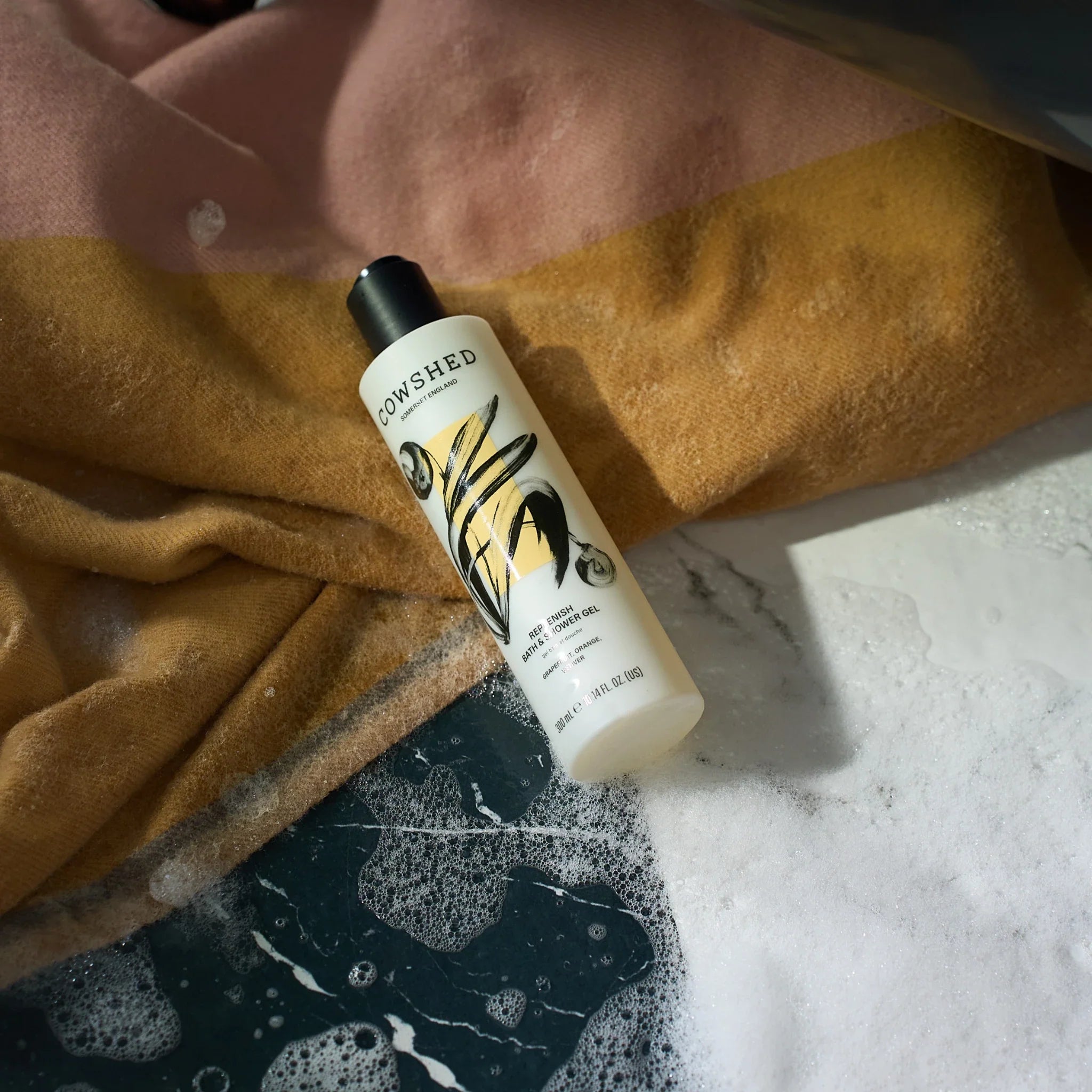 Cowshed replenish shower gel on the floor of a bathroom with bubbles and a towel