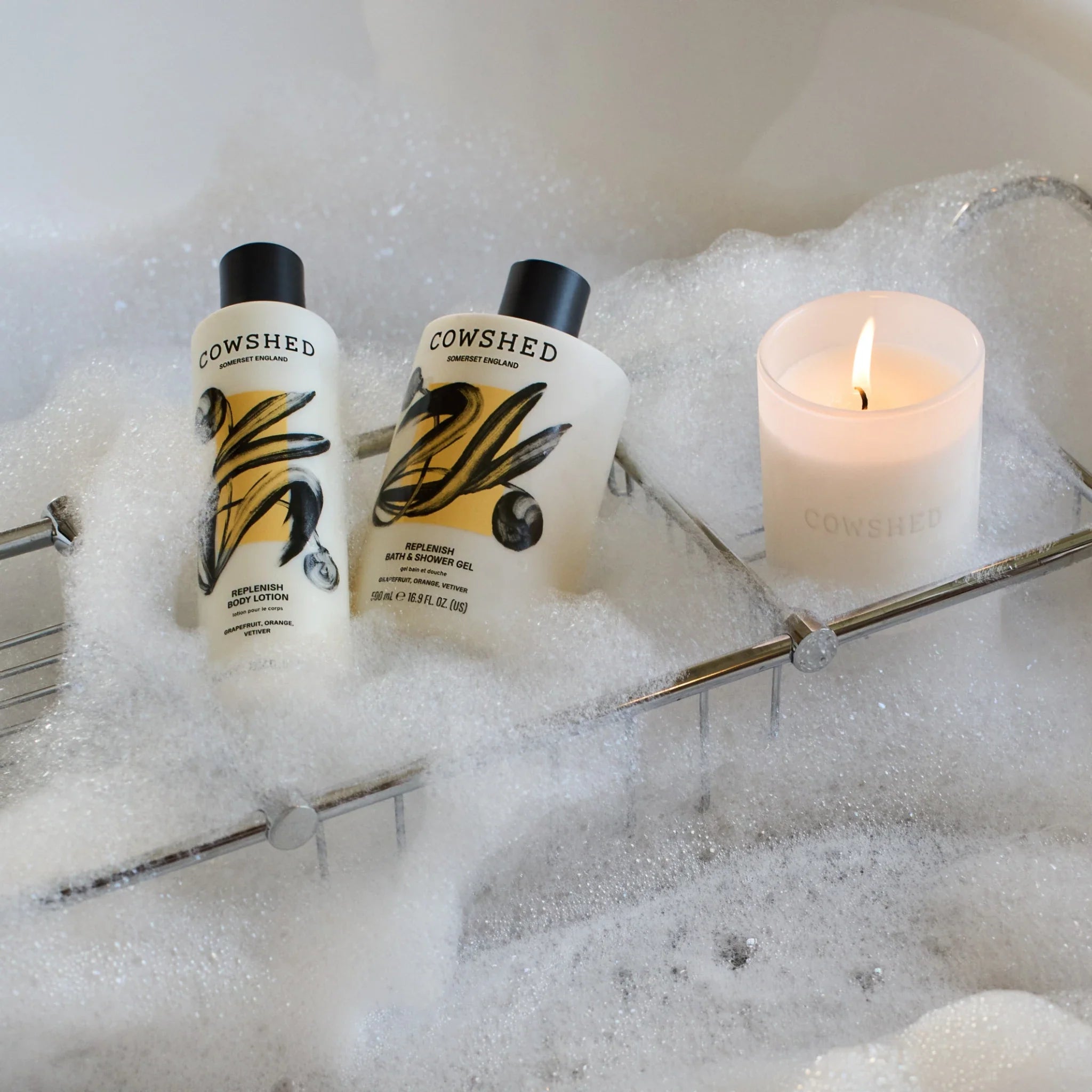 Cowshed replenish bath and body set in a bubble bath with a lit candle on a rack