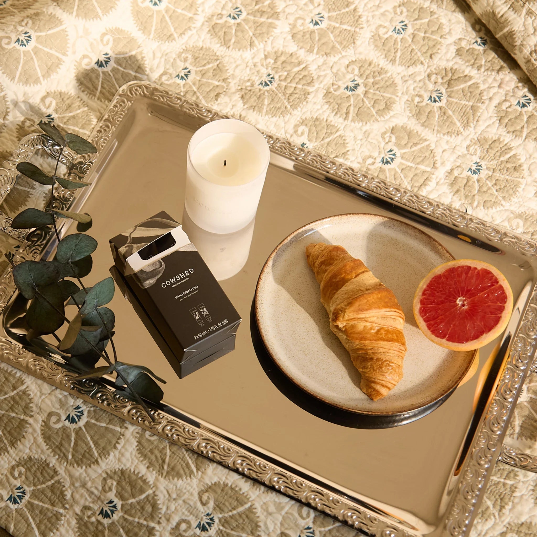 Silver tray with a croissant, grapefruit, a candle & hand cream duo on patterned bedding.