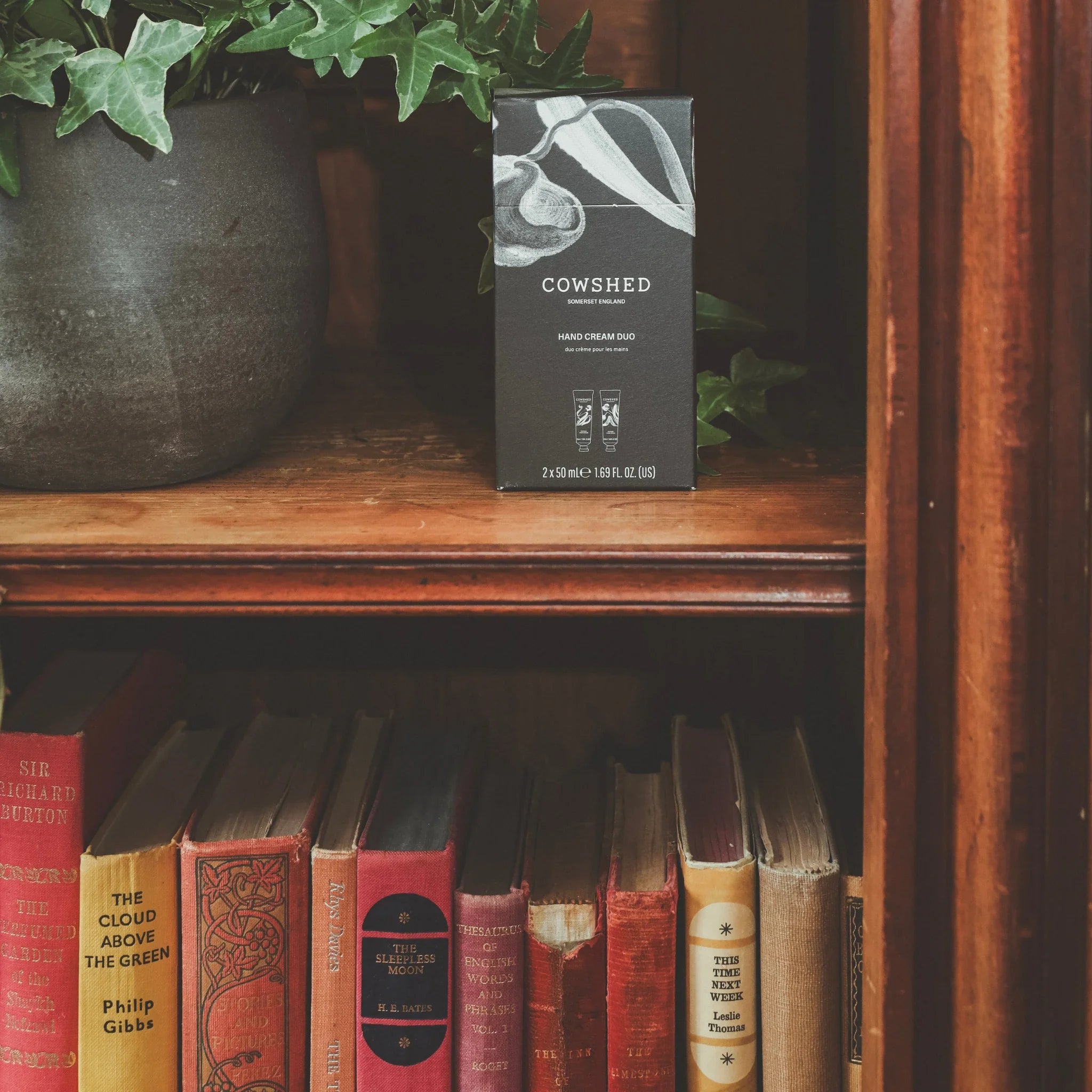 Cowshed hand cream duo on a wooden bookshelf with a plant