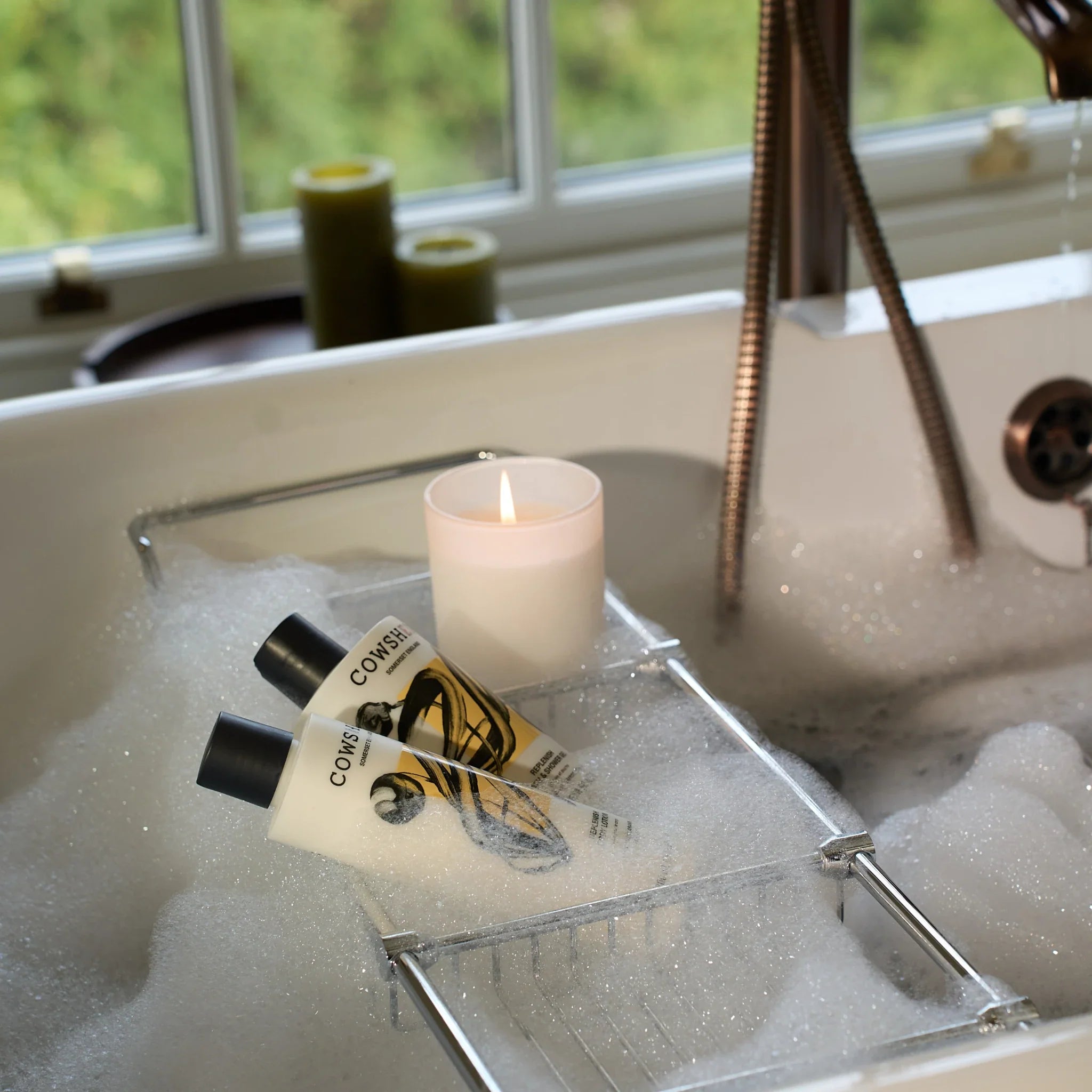 Bottles of Cowshed soap and lotion and a lit candle with a bubble bath.