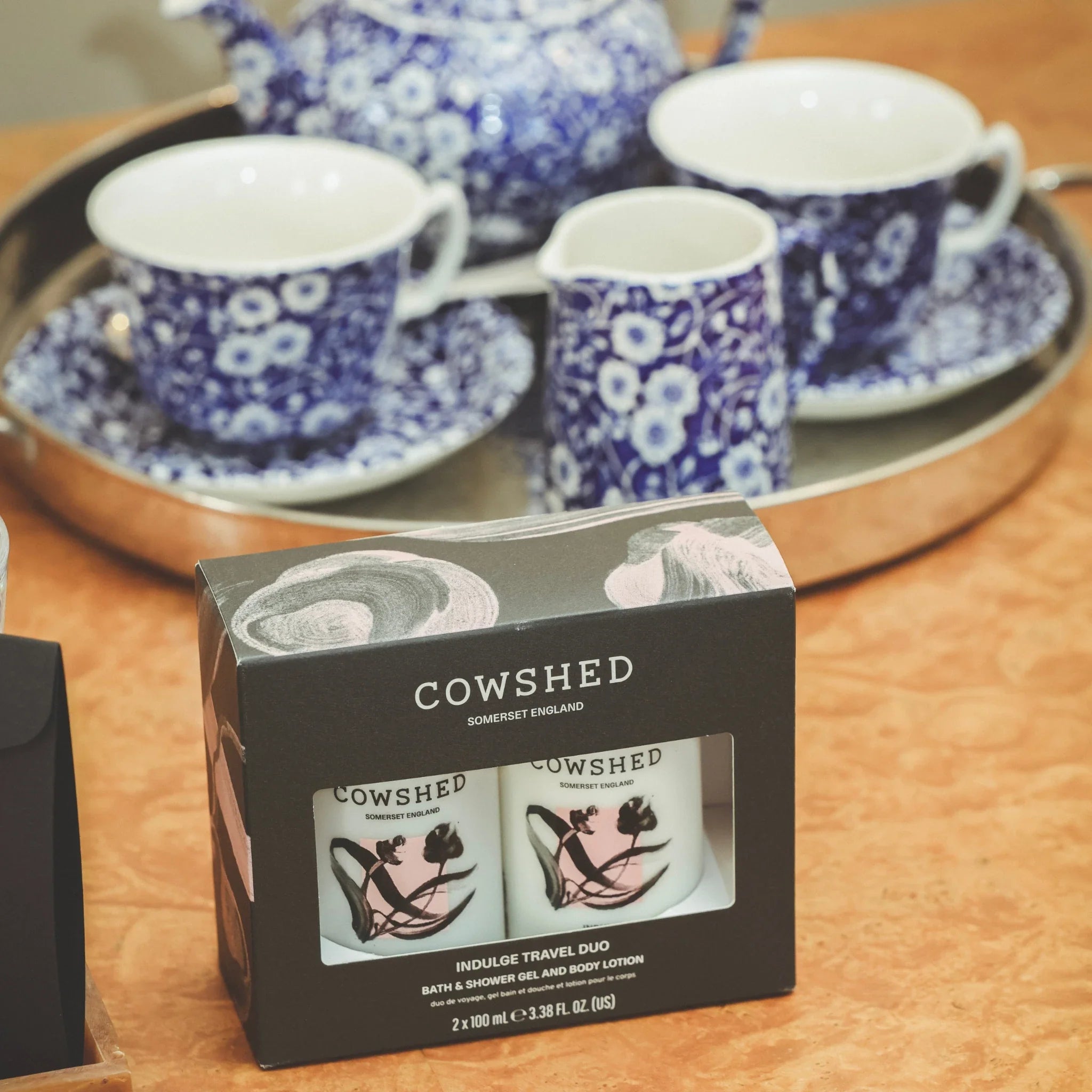 Cowshed Indulge product packaging on a wooden surface with floral teacups in the background