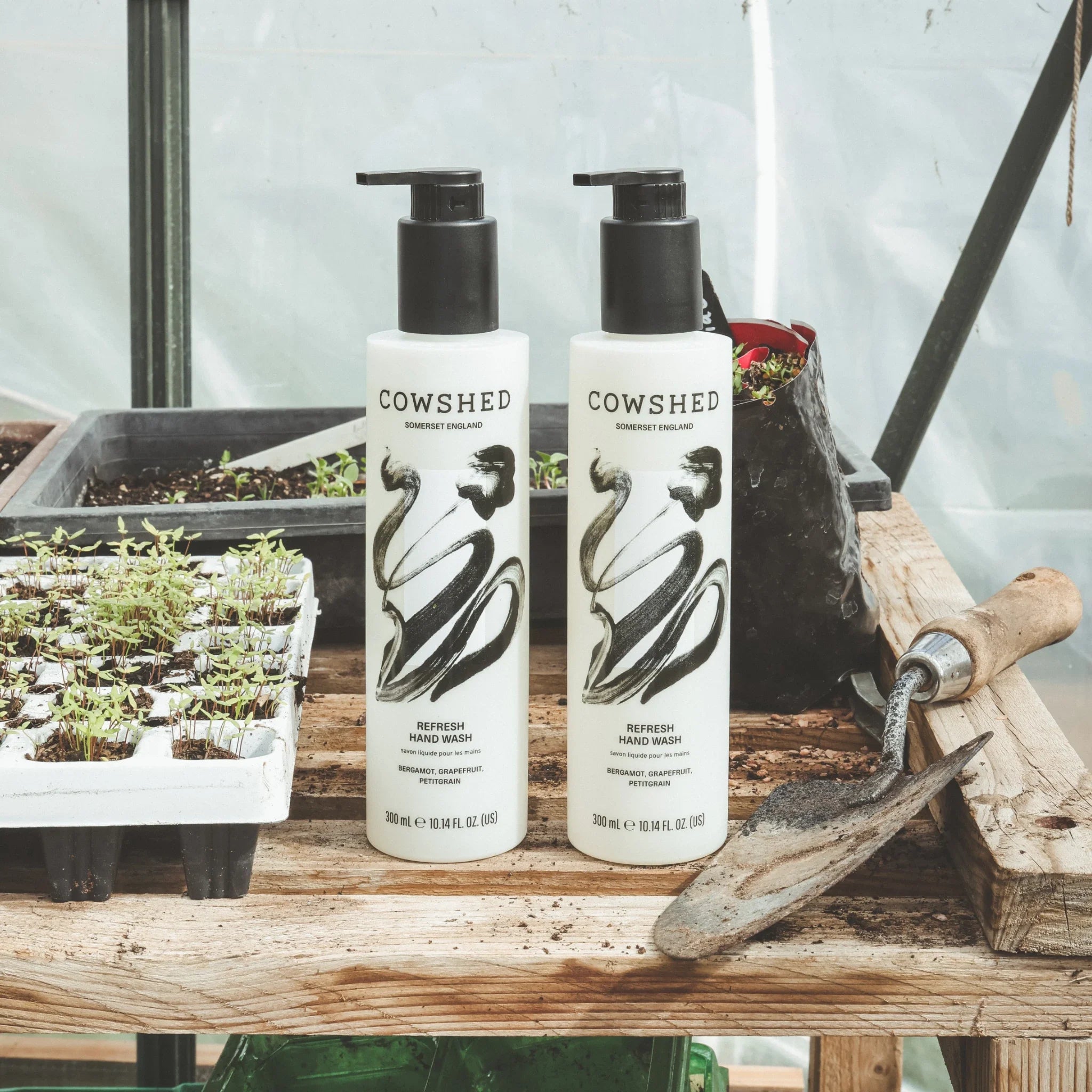 Two Cowshed hand wash bottles on a wooden bench with gardening tools in the background.