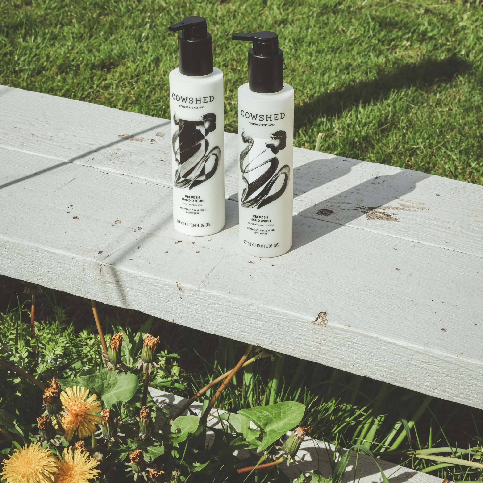 Two Cowshed hand care products on a wooden bench with grass and flowers in the background