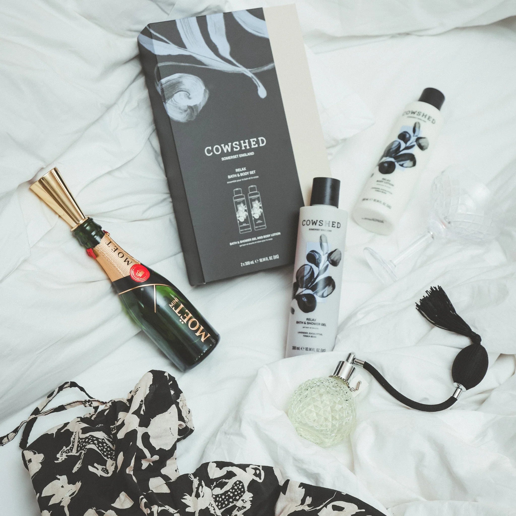 Cowshed Relax product set displayed with a champagne & perfume bottle on white linen bedding