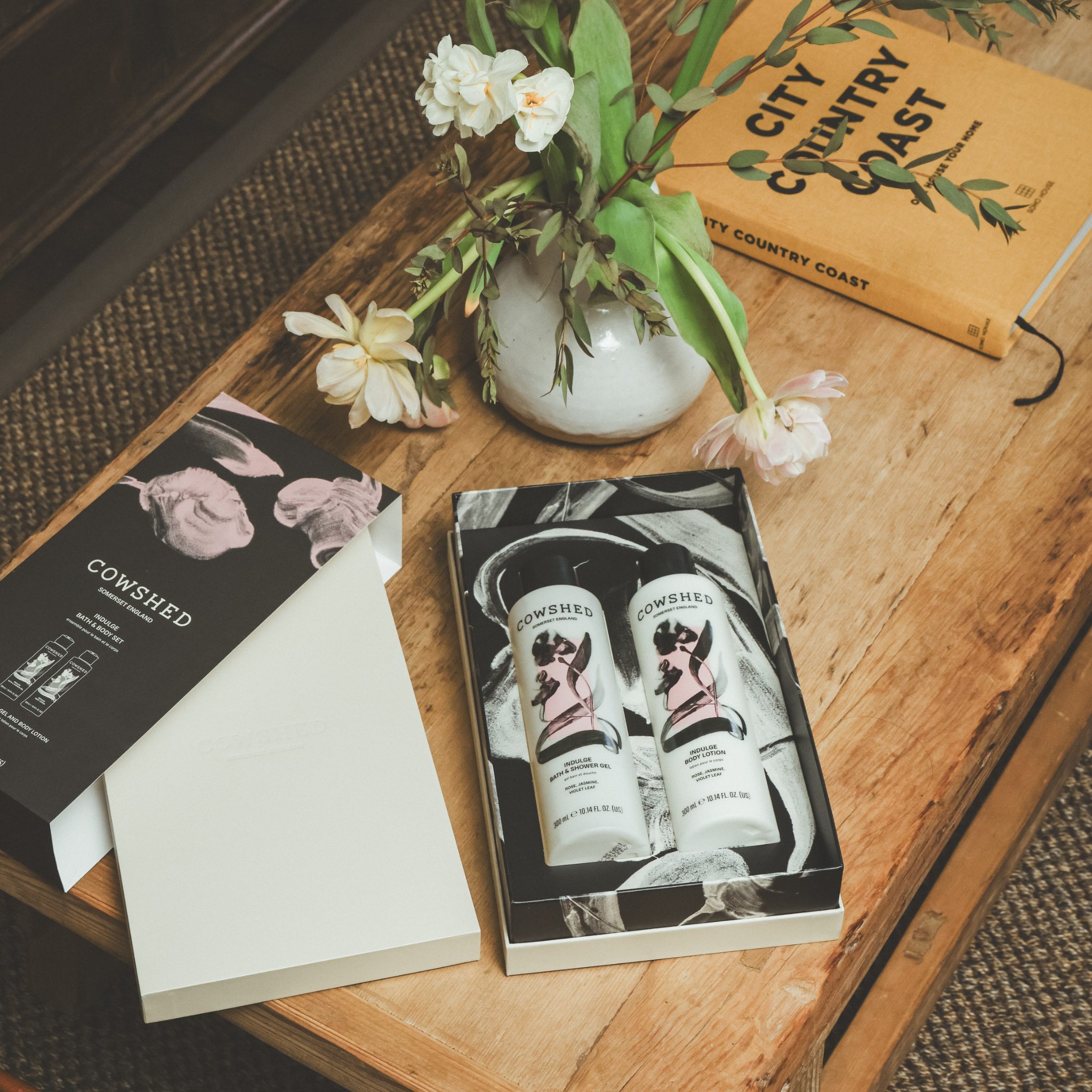 Cowshed products on a wooden table with flowers & a book in the background