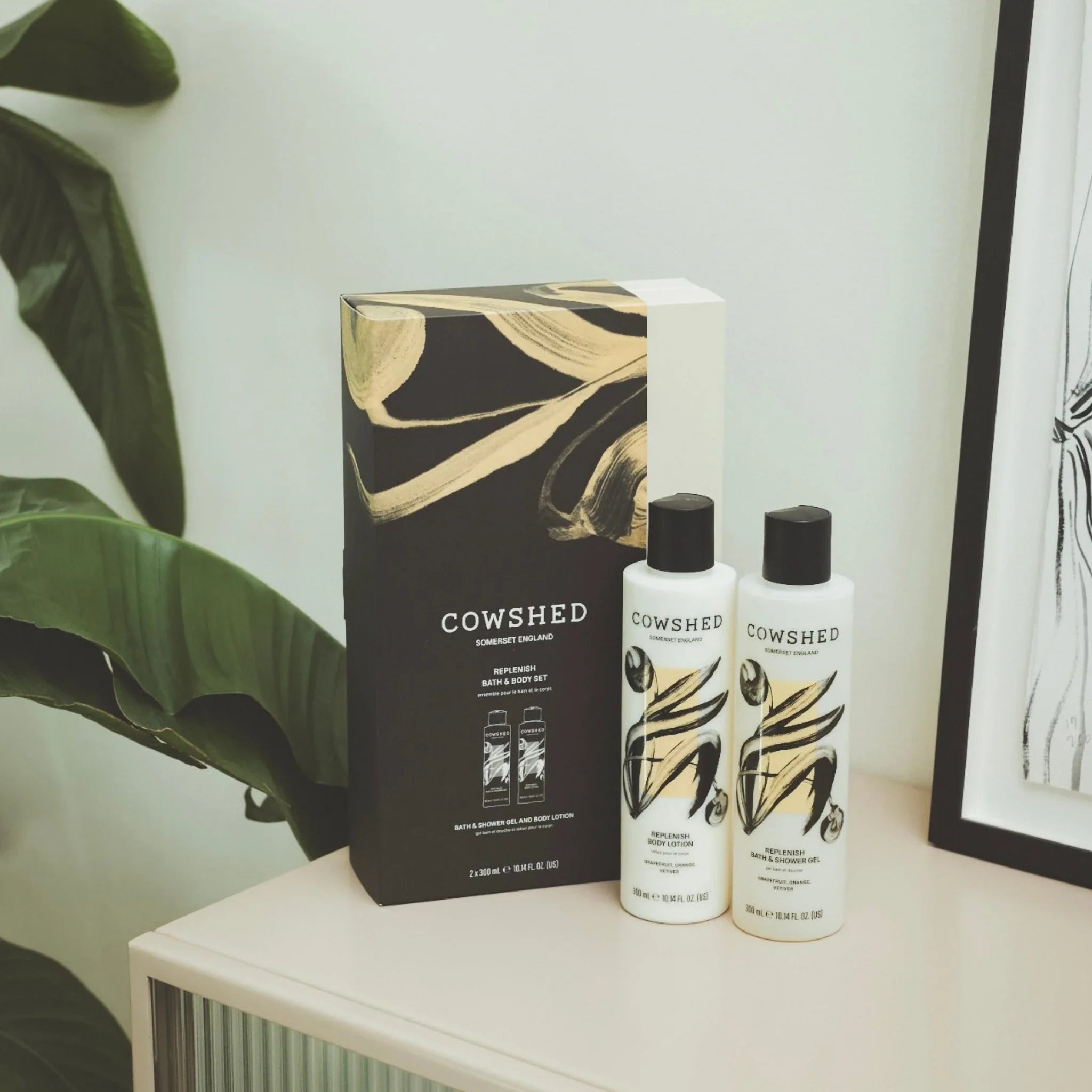 Cowshed Replenish bath and body duo on a white surface with a plant and framed picture in the background