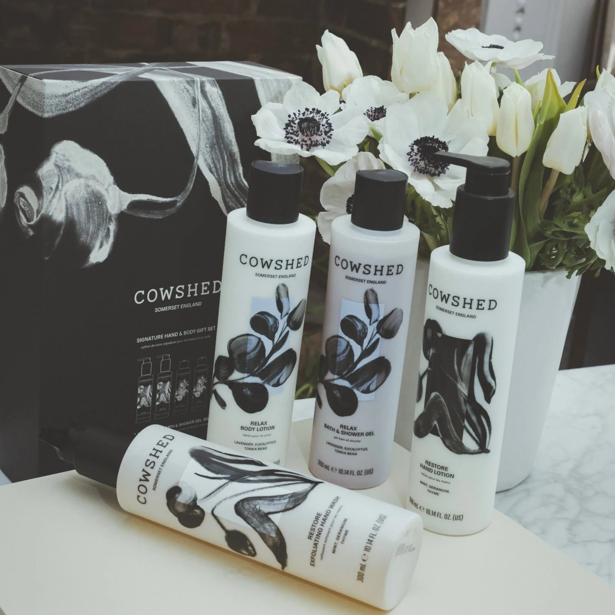 Cowshed skincare products on a table with a decorative box and flowers in the background