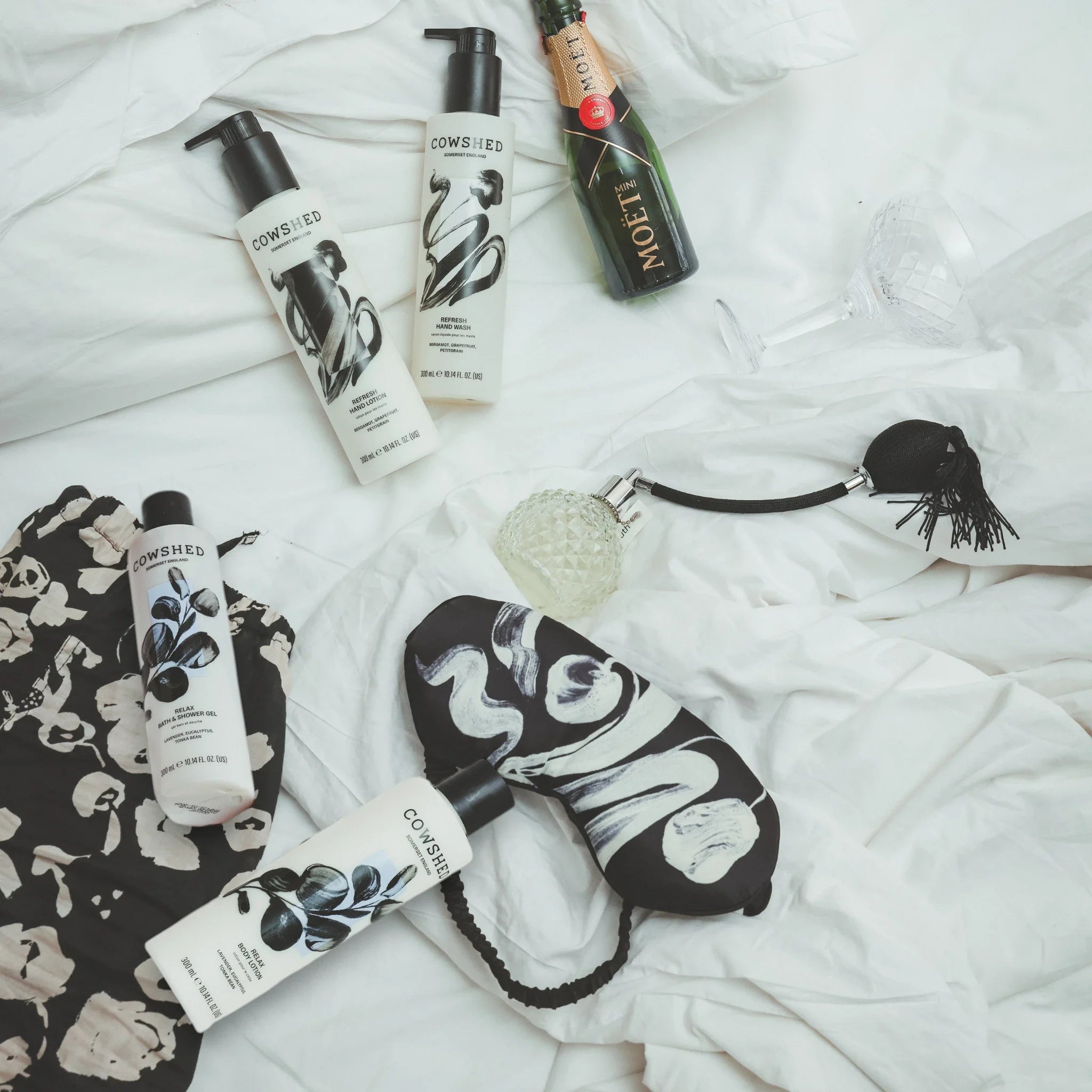 Collection of Cowshed products including a sleep mask & perfume bottle on a white duvet