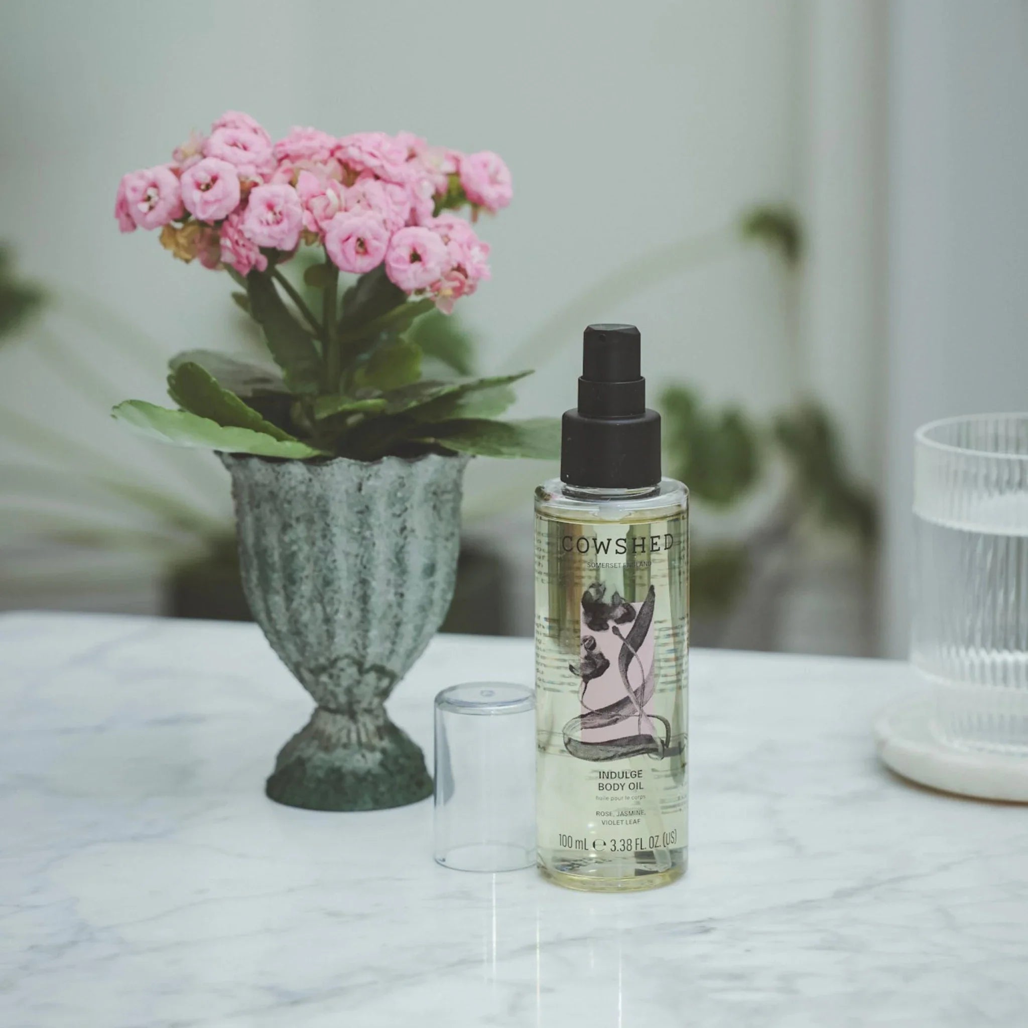 Cowshed Indulge body oil product on a marble surface with pink flowers in the background