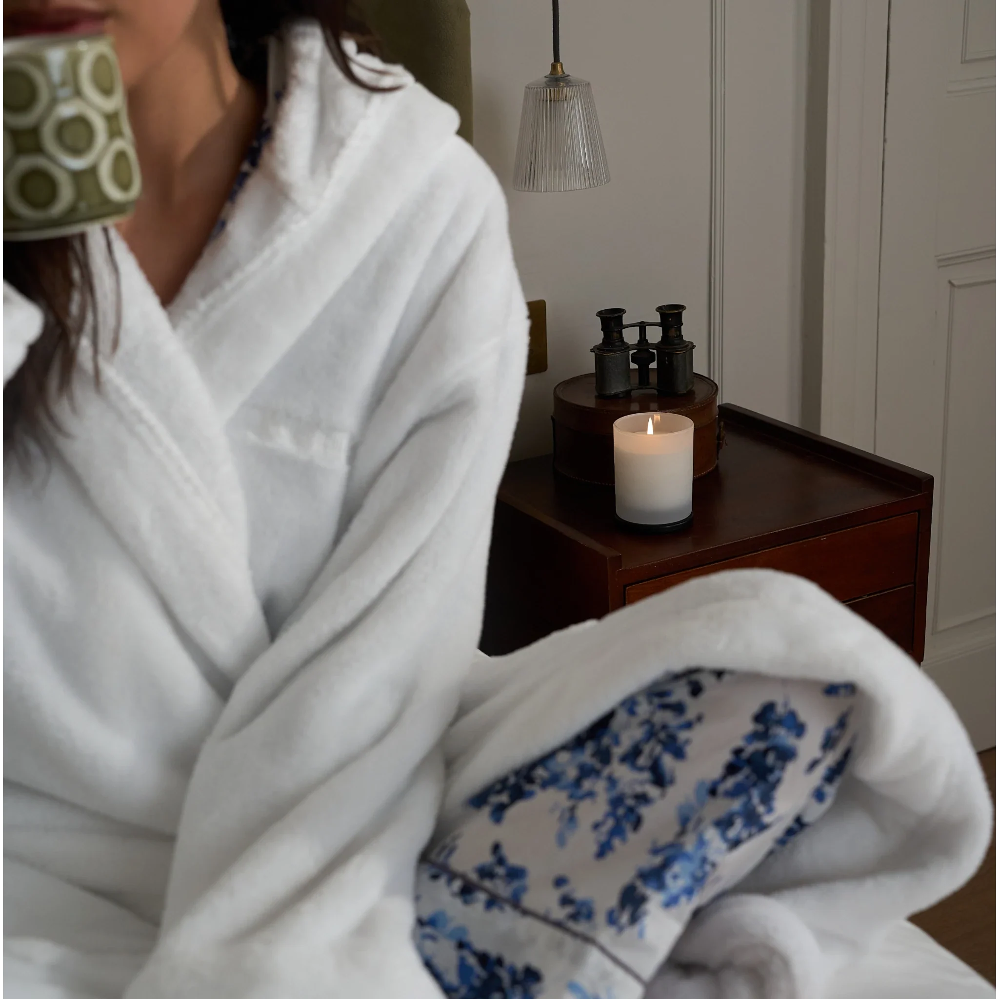 Person in a white robe holding a mug, sitting by a candlelit nightstand.
