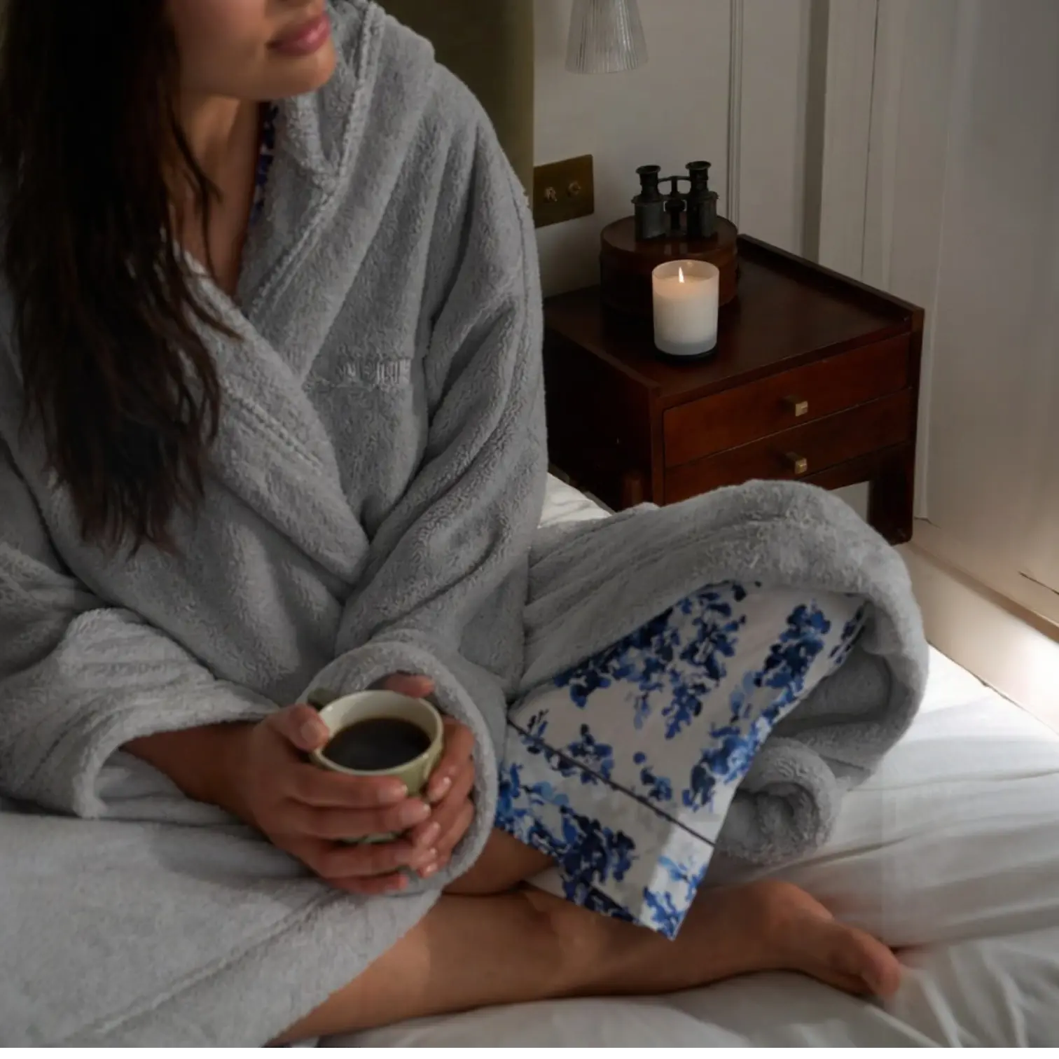Person in a gray robe holding a cup, sitting on a bed with a floral blanket.