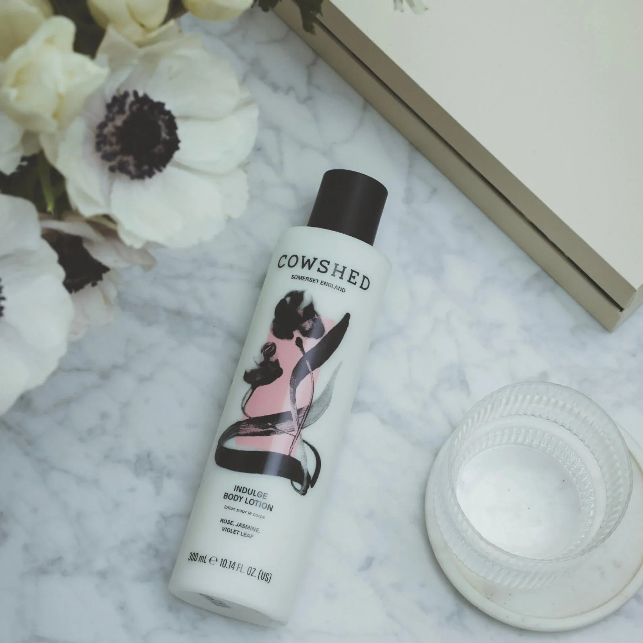 Cowshed body lotion bottle on a marble surface with flowers and a book in the background