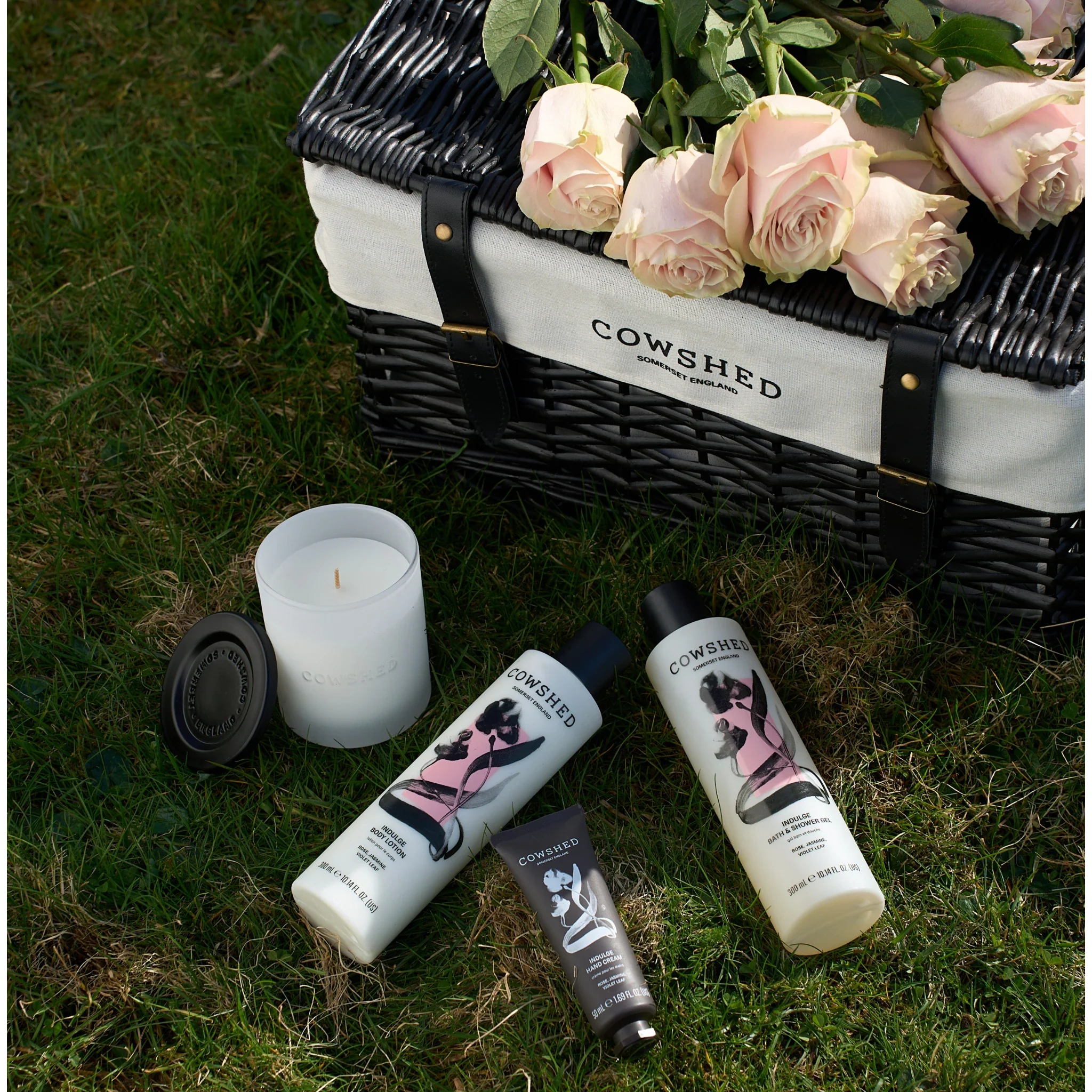 Cowshed Indulge range on the grass next to a hamper and roses