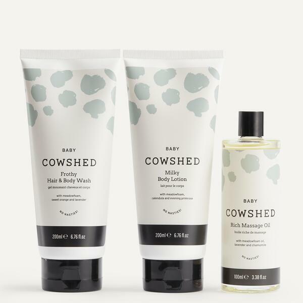 A set of three baby care products by Cowshed, including hair and body wash, body lotion, and rich massage oil, all packaged in white bottles with green leaf patterns.