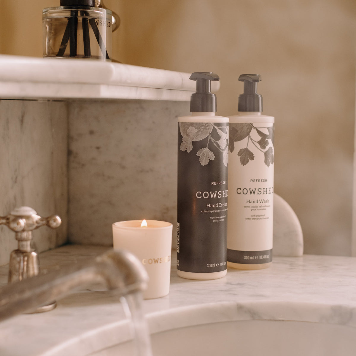 Two bottles of Cowshed refresh hand lotion and hand wash on a marble surface with a lit candle.
