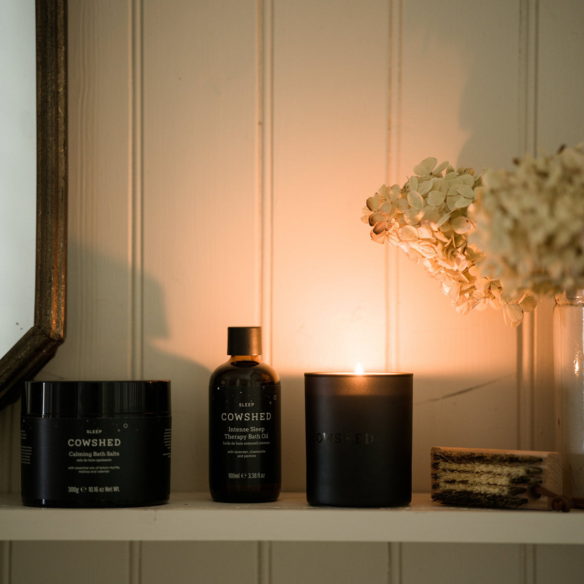 Cowshed products including a jar, bottle, and candle on a shelf with a soft light background.